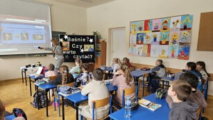 Widok na klasę siedzącą w ławkach, przed nimi nauczycielka pokazuje na tablicy baśnie, obok stoi tablica z pytaniami. Baśnie? Czy warto czytać?