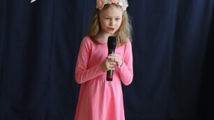 Uczennica ubrana w różową sukienkę śpiewa piosenkę.