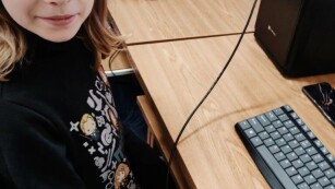 Dziewczynka siedzi przy stanowisku komputerowym i trzyma kabelek od mikrokontrolera Arduino