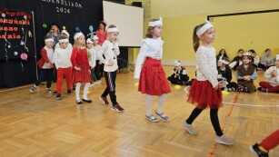 Grupa przedszkolaków ubrana na biało czerwono z opaskami na głowie w kształcie Koziołka Matołka idzie gęsiego przez salę gimnastyczną.