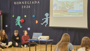Na zdjęciu widać rzutnik z ekranem, na którym prezentowane są pytania do konkursu, z boku uczniowie przyglądają się uważnie w celu udzielenia poprawnej odpowiedzi.