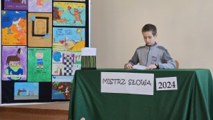 Na pierwszym planie uczeń siedzi za stolikiem nakrytym zielonym suknem i głośno czyta książkę. Obok niego stoi tablica z plakatami przygotowanymi przez uczniów prezentującymi sceny z książek Makuszyńskiego.