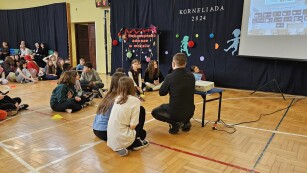 Uczniowie siędzą na podłodze w sali gimnastycznej, słuchają nauczyciela, który tłumaczy zasady konkursu o życiu i twórczości Kornela Makuszyńskiego.  Z prawej widoczny ekran z rzutnikiem. Z lewej pozostali uczniowie, którzy kibicują.