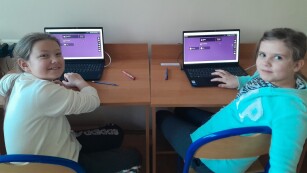 Dwie uczennice przy laptopach, odwrócone do obiektywy uśmiechają się.