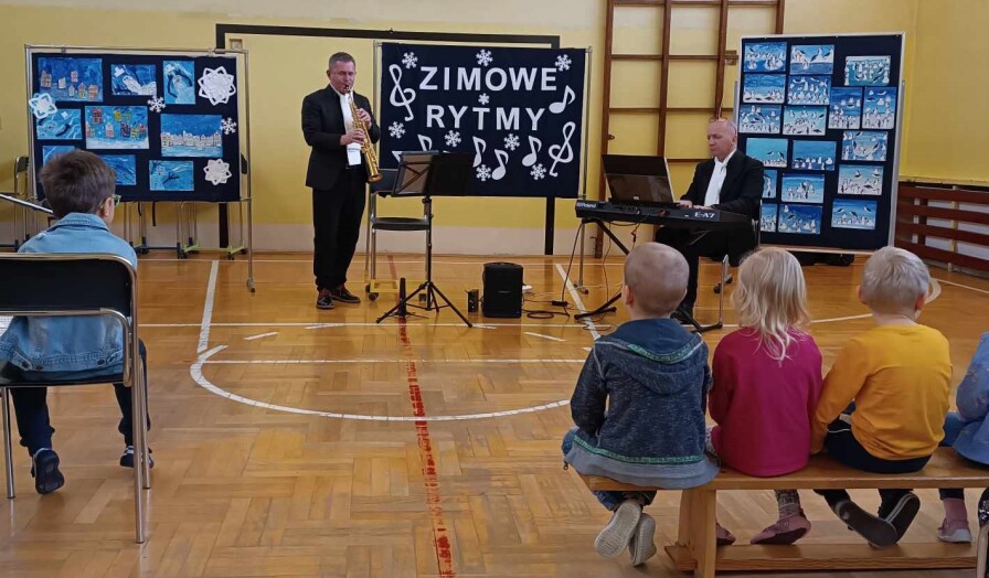 Dwóch muzyków gra na instrumentach, uczniowie na krzesełkach przysłuchują się.